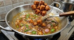 Gently lowering golden-brown fried chicken pieces into the bubbling, simmering dark red Manchurian gravy, surrounded by chunky bell peppers and onions in a hot wok."