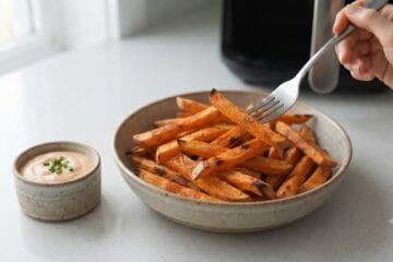 Golden brown and crispy Air Fryer Sweet Potato Fries served hot in a rustic bowl with a side of creamy dipping sauce, garnished with chives on a clean kitchen counter."