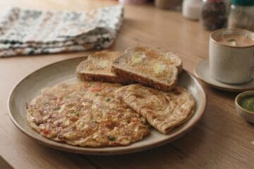 Ek bhuni hui, gold-brown Masala Omelette, chopped pyaz, tamatar, aur hari mirch ke sath, ek rustic ceramic plate par parosi gayi hai. Saath mein garam, folded Lachha Paratha aur do slices buttered multi-grain toast rakhe hain. Side mein ek choti katori mein hara dhaniya-pudine ki chutney hai, aur background mein ek cup bhap lete hue Masala Chai ka hai. Sab kuch ek warm wooden table par hai.