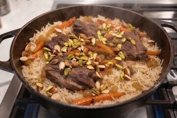 A final serving presentation of Afghan Kabuli Pulao in a cast-iron pot featuring fluffy rice topped with tender mutton pieces, caramelized carrots, raisins, and garnished with toasted almonds and pistachios.