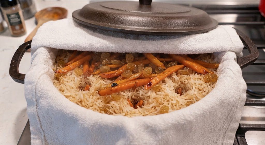 Basmati rice boiling in a large cast-iron pot with rich meat broth, sprinkled with garam masala on top, while cooking an Authentic Afghan Kabuli Pulao Recipe.