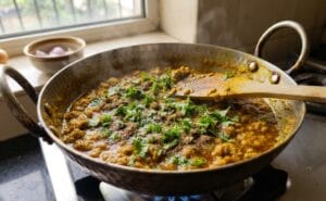 A close-up photograph of the finished chicken keema in the hammered iron karhai from image_16.png, now garnished with a generous sprinkling of fresh chopped green coriander and garam masala powder. A wooden spoon is resting in the dish, which is steaming gently, indicating it's ready for this final step of a traditional mutton Keema Recipe. The background kitchen counter and window view are softly blurred