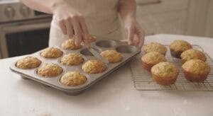 A cooling rack holding freshly baked muffins next to a metal muffin tray where a hand uses a butter knife to gently loosen a golden-brown, baked Cheesy Zucchini Muffins from its mold.