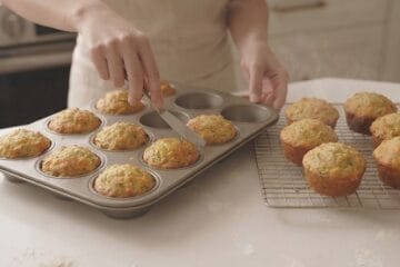 A cooling rack holding freshly baked muffins next to a metal muffin tray where a hand uses a butter knife to gently loosen a golden-brown, baked Cheesy Zucchini Muffins from its mold.